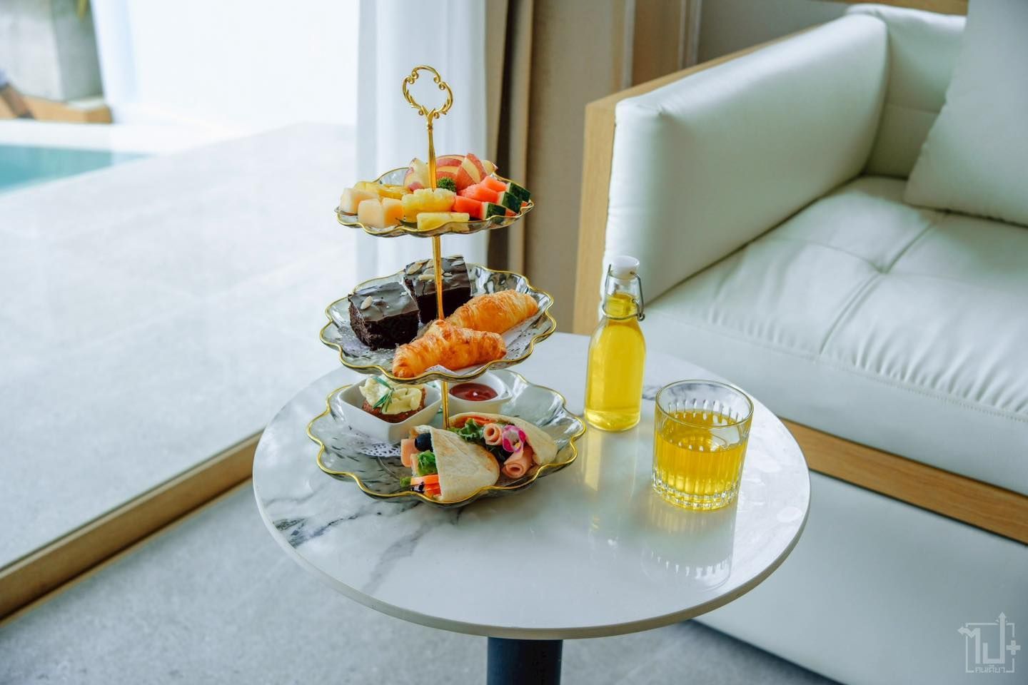 Three-tiered dessert tray with various pastries and fruit on a table with a drink and a pool view.