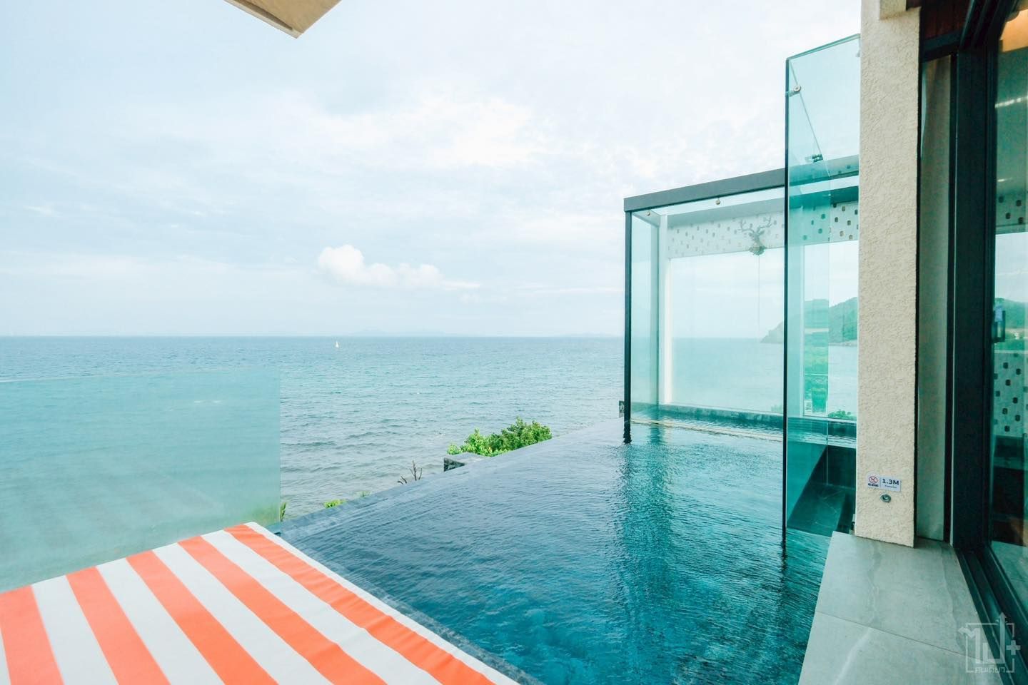 Infinity pool overlooking the ocean, modern building with glass walls, striped awning, overcast sky.