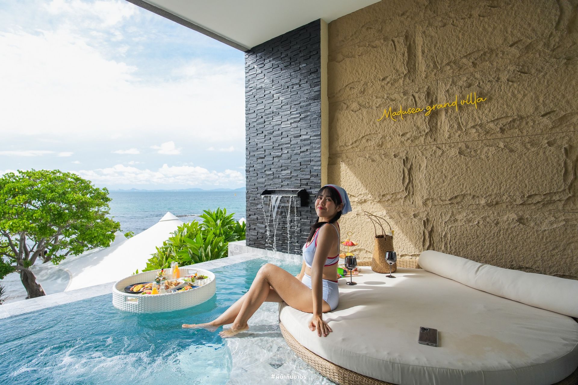 Woman in swimsuit sits by a pool overlooking the ocean. Floating tray with food, textured wall, water feature.