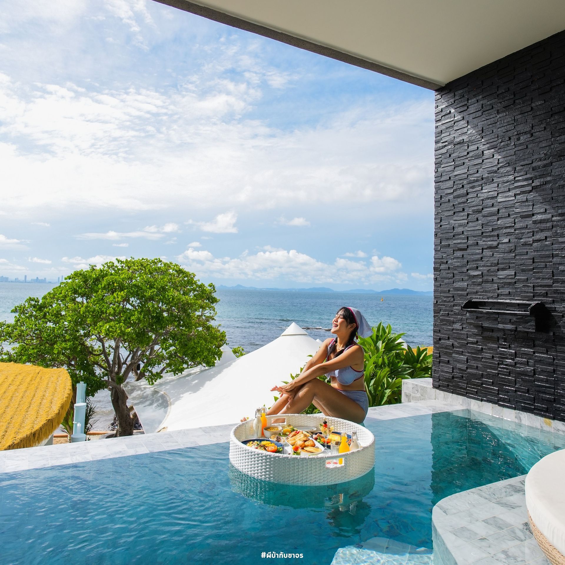 Woman in pool with floating breakfast tray overlooking the ocean. Sunny day.