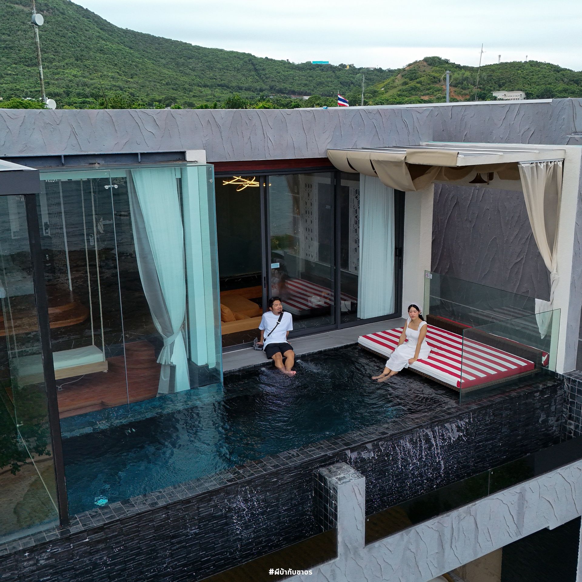 Rooftop pool with two people. One sits in the water, the other on a daybed. Mountains in the background.