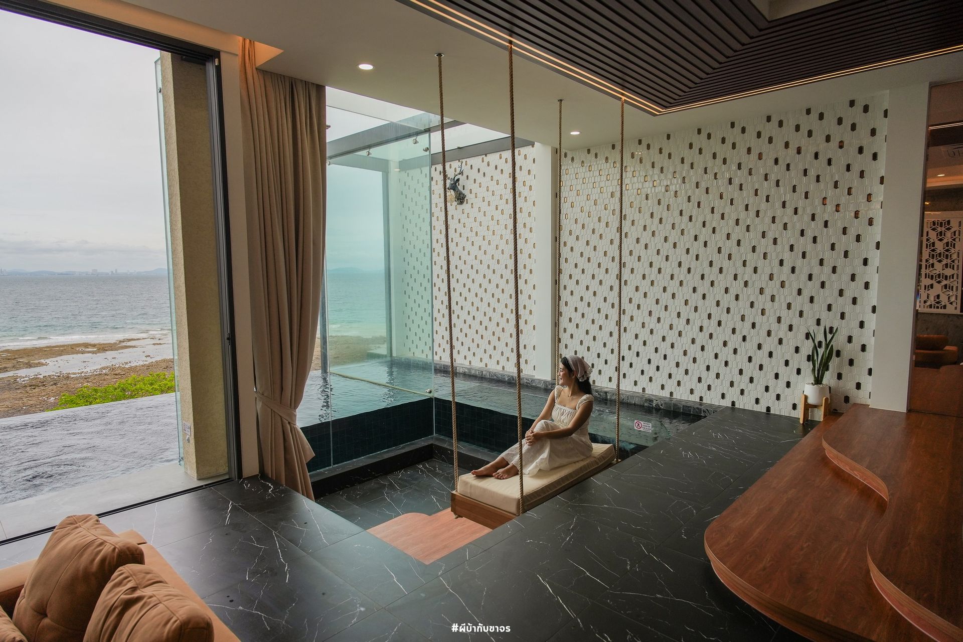 Woman sitting on swing indoors, overlooking ocean view through glass wall.
