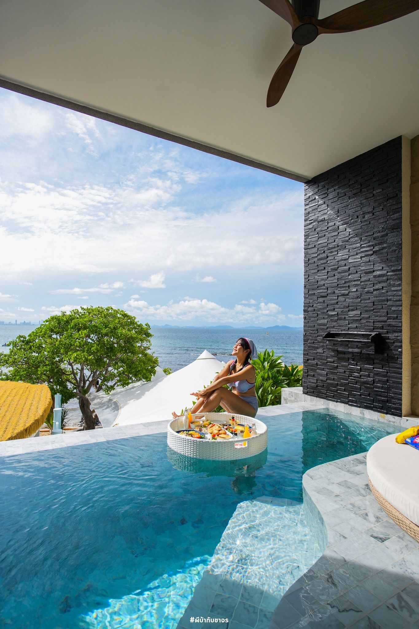 Woman relaxing in a pool with food tray, overlooking ocean and blue sky.