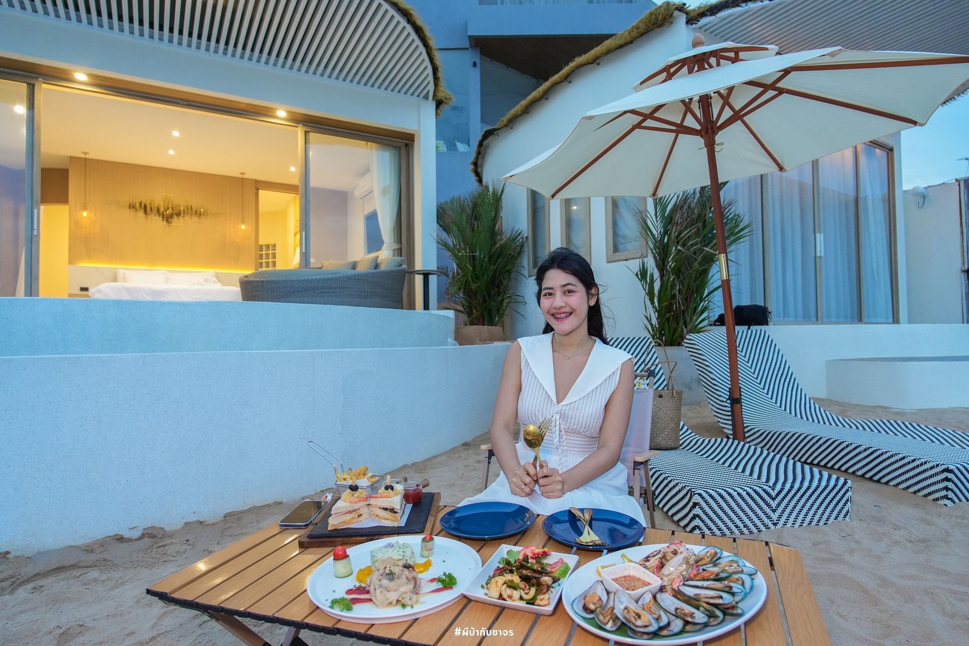 Woman in white dress at table with food on beach, by white building, holding drink.