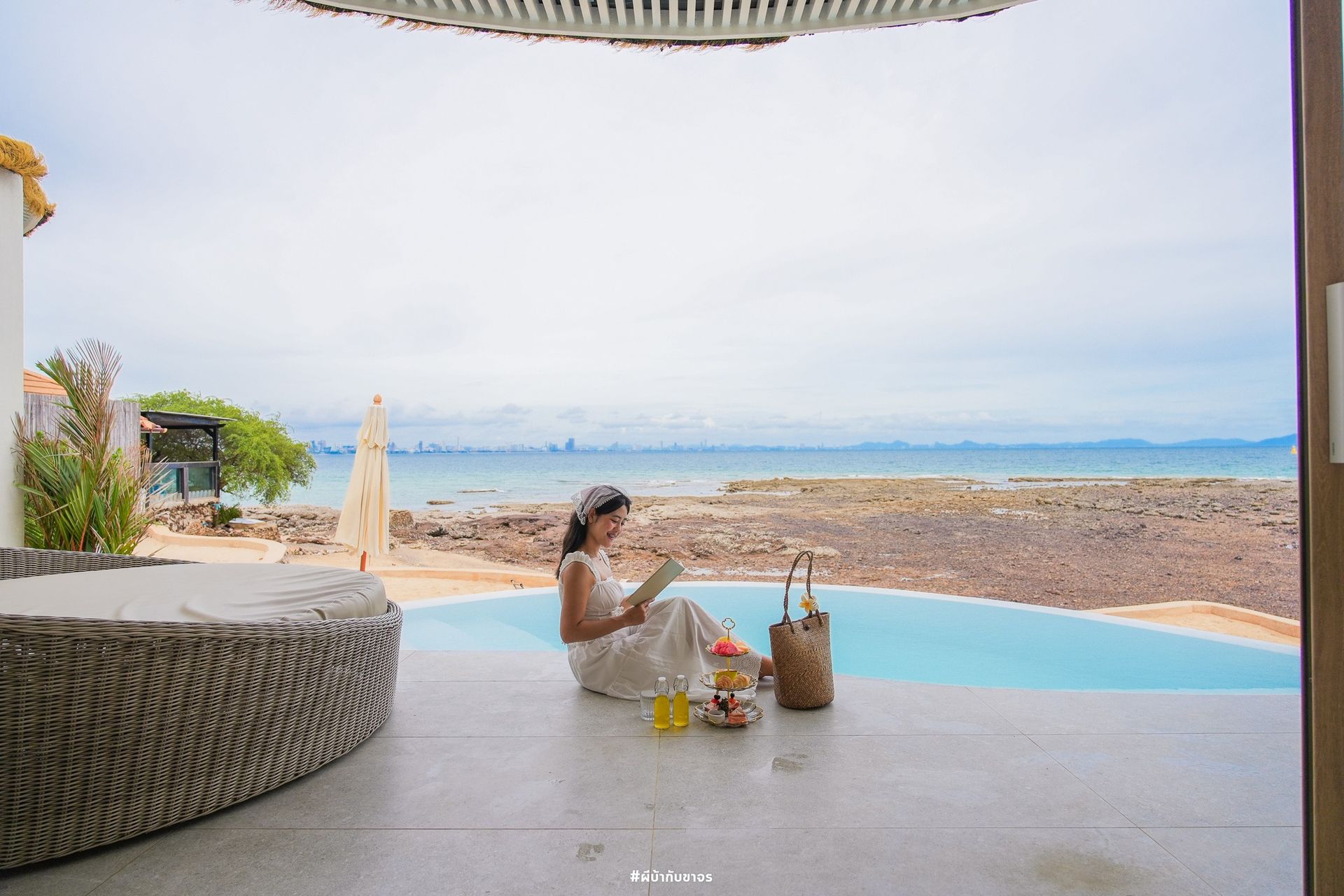 Woman reading by a pool overlooking the ocean. She is seated on a patio, with a book and food nearby.