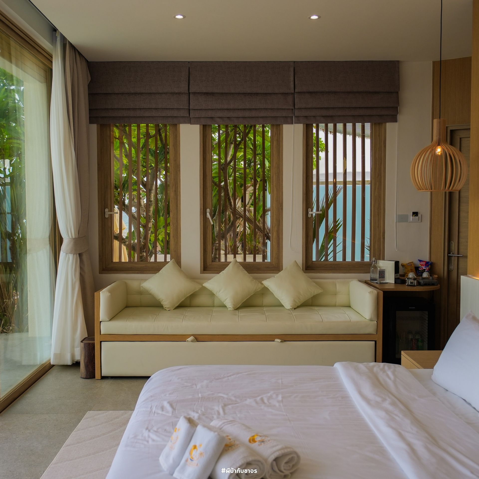 Cozy hotel room with cream-colored bed, couch, and window with vertical blinds, view of greenery.