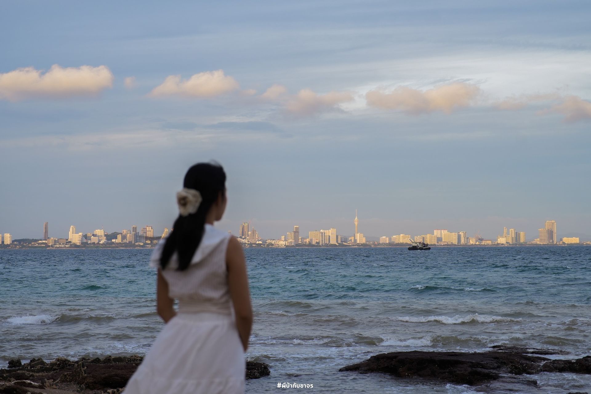 Woman in white dress looks out at city skyline over ocean under a cloudy sky.