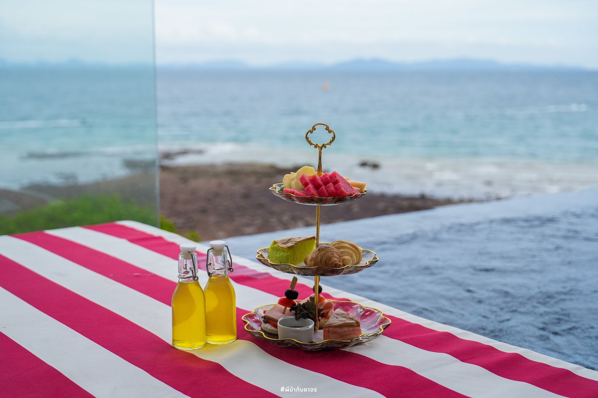 Tiered fruit and pastries on red and white striped cloth, with ocean view and two bottles of yellow liquid.