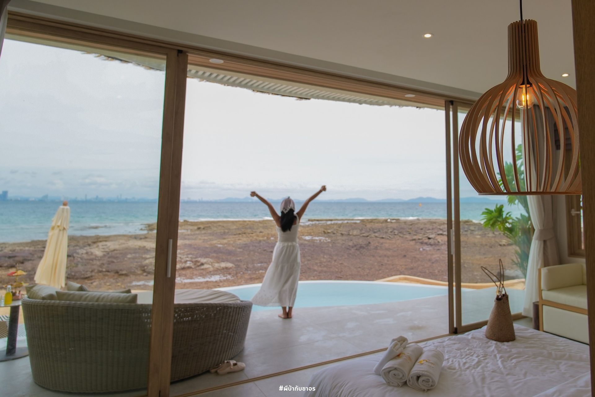 Woman in white outfit stretches towards ocean view from a room with large glass doors.