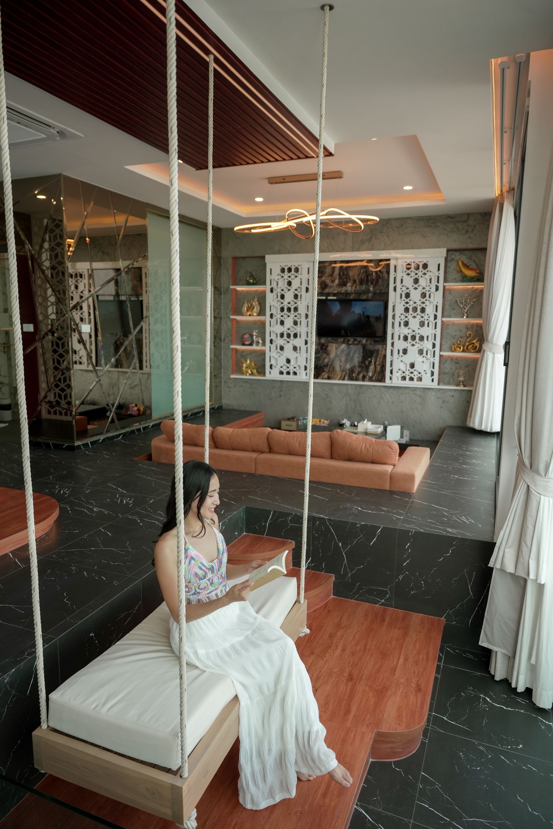 Woman on a swing in a modern living room, reading. Orange accents, large windows, and a sofa visible.