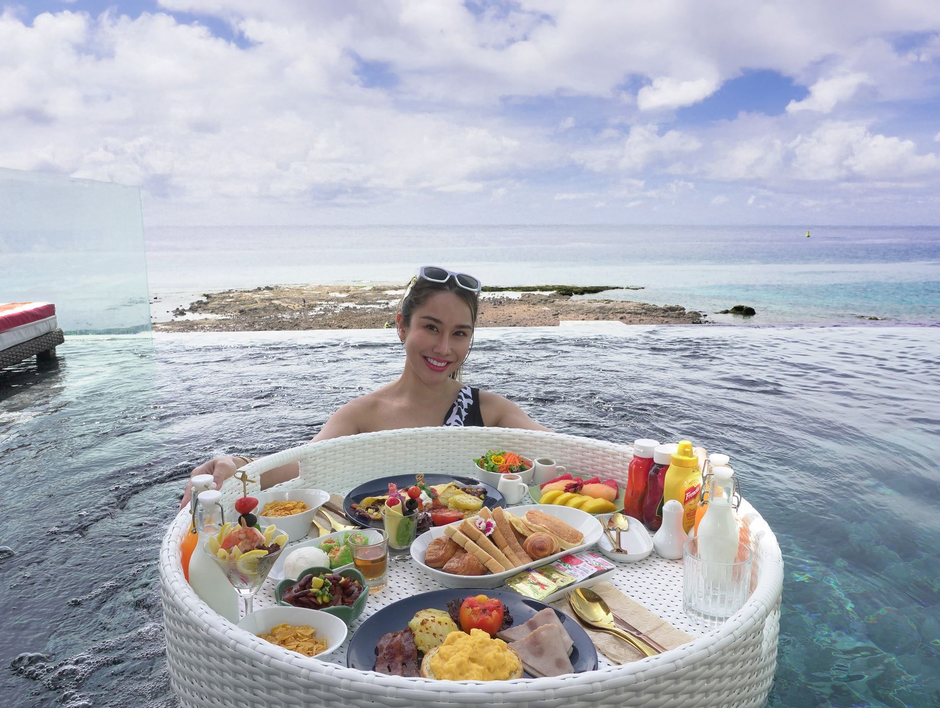 Woman in pool holding a floating tray of food; ocean view with blue sky.