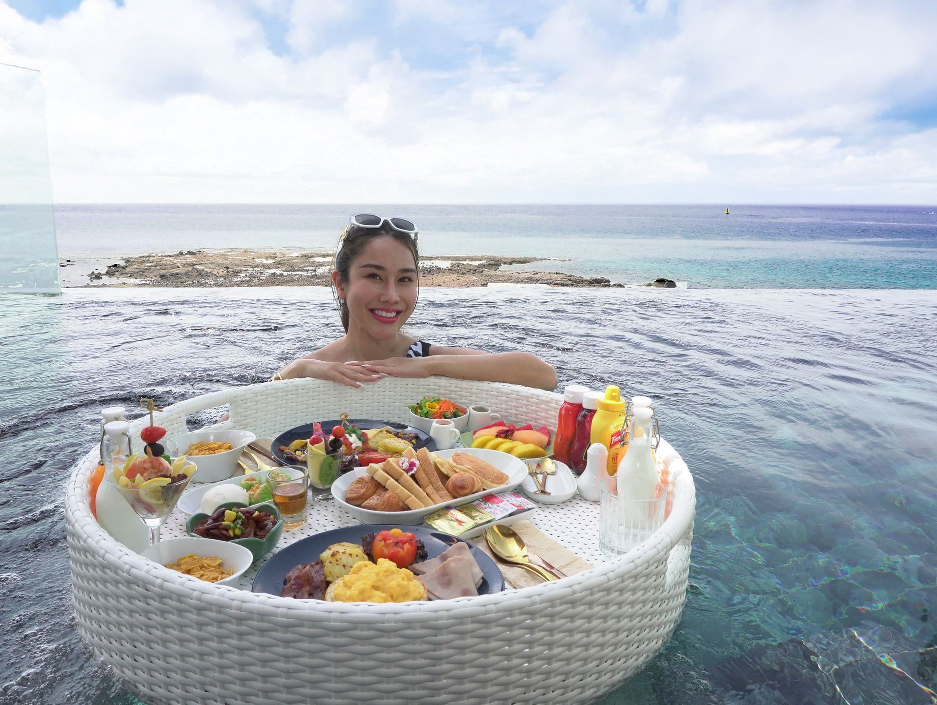 Woman in pool with floating breakfast tray, ocean backdrop. Smiling, sunny day.