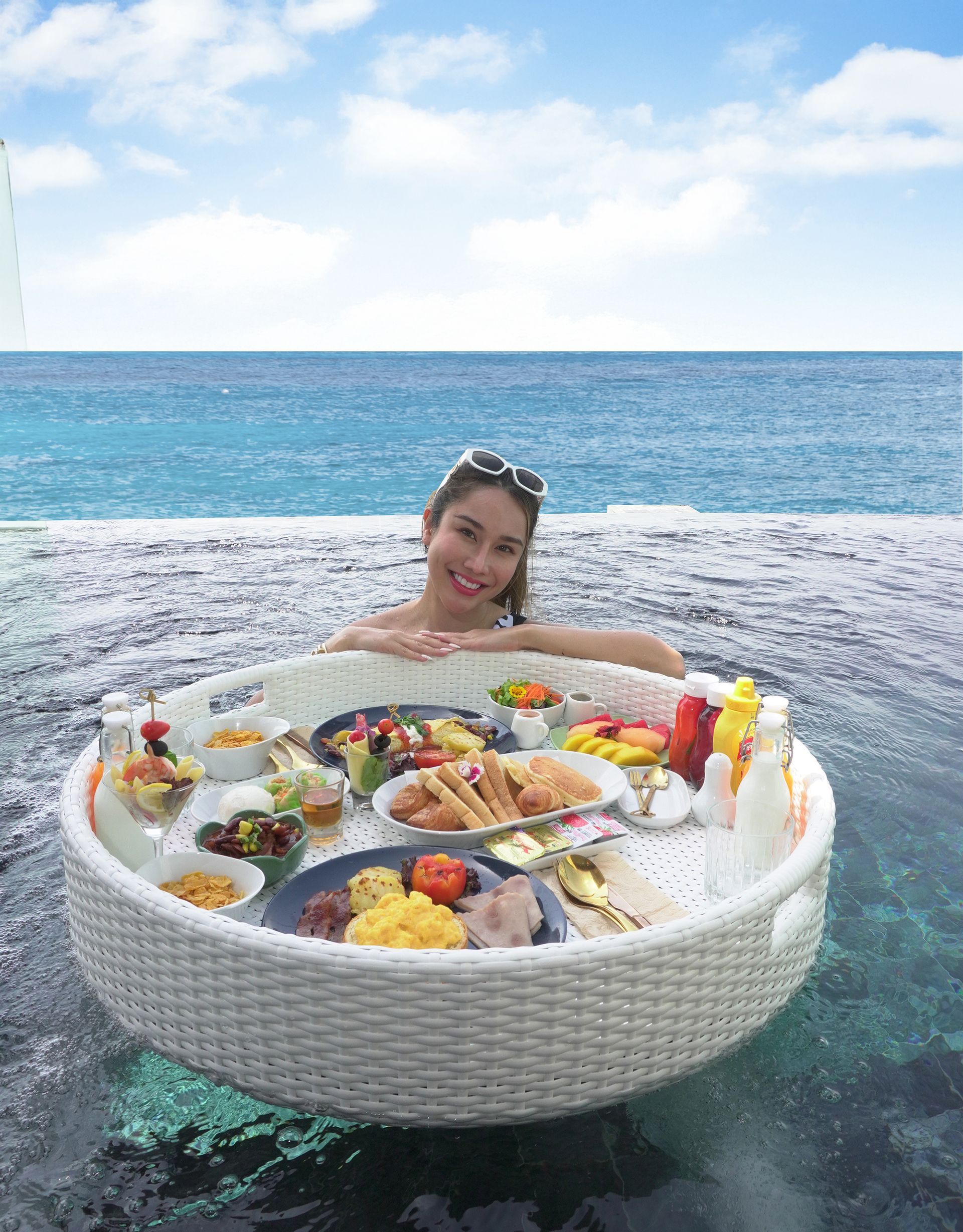 Woman in a pool enjoying a floating breakfast tray with various foods, sunny day, ocean backdrop.