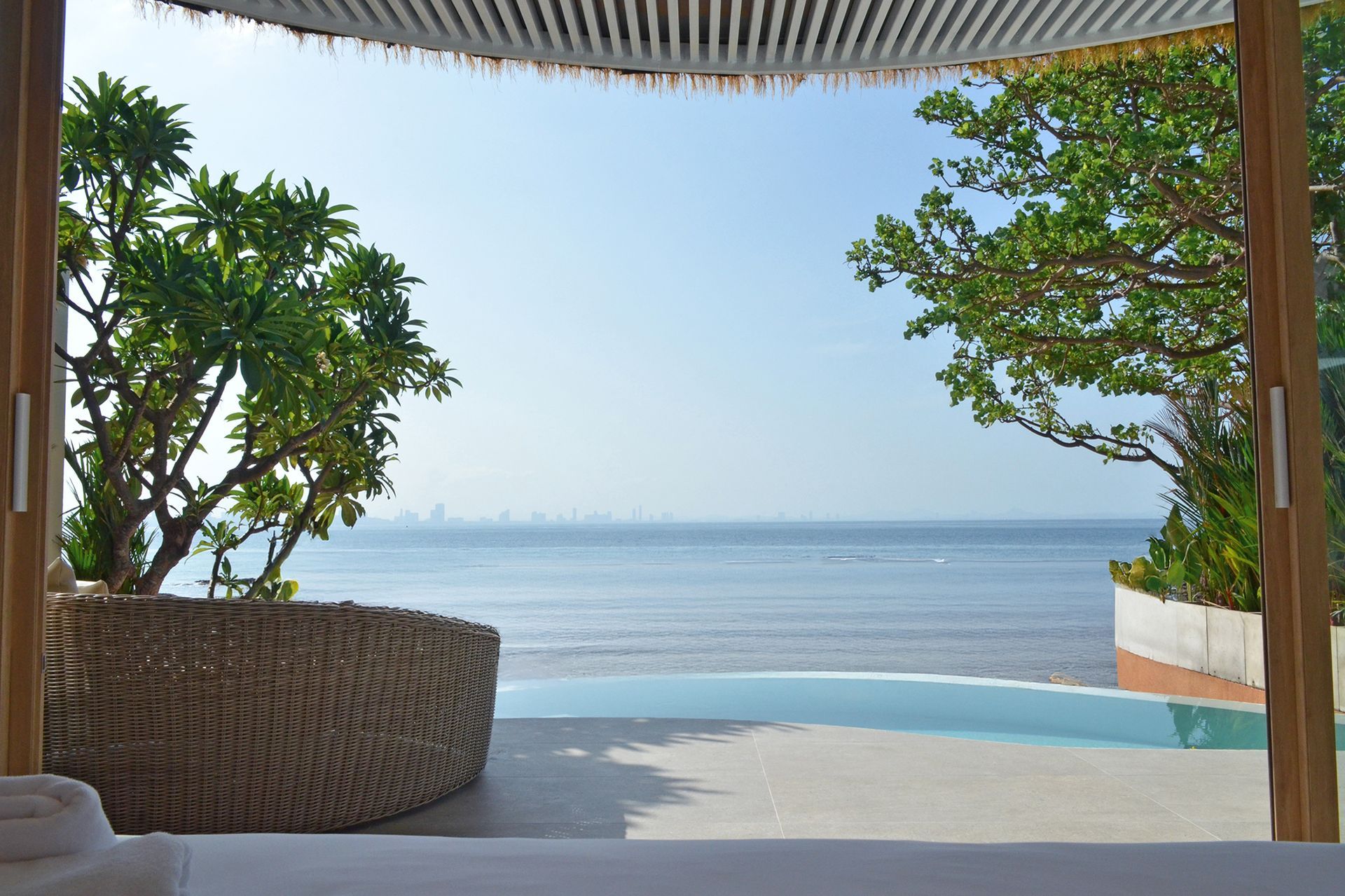 View of ocean from a covered lounge area with trees, a pool, and city in the distance.