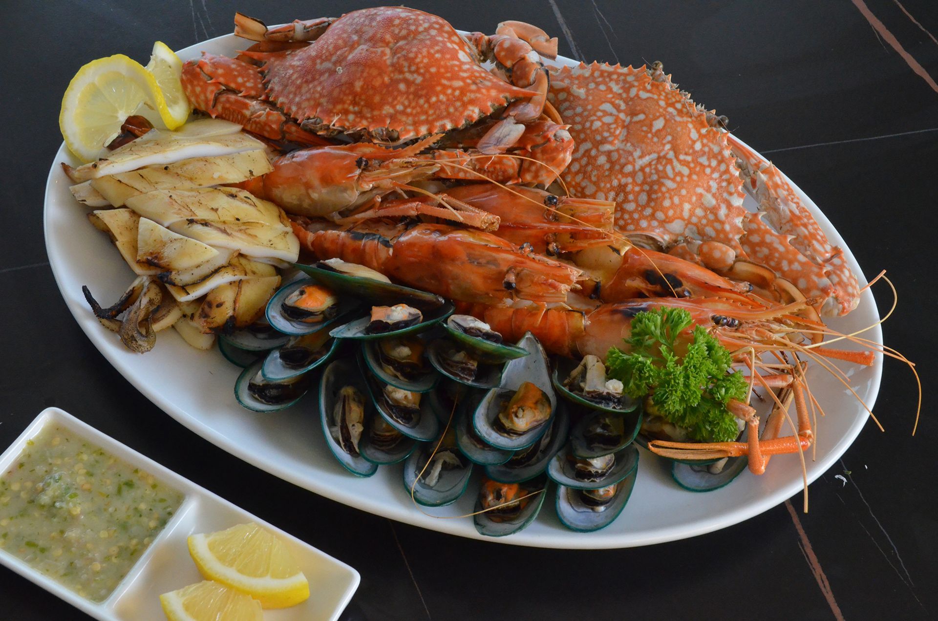 Seafood platter: crab, shrimp, mussels, grilled squid, lemon slices, and dipping sauce on a white plate.