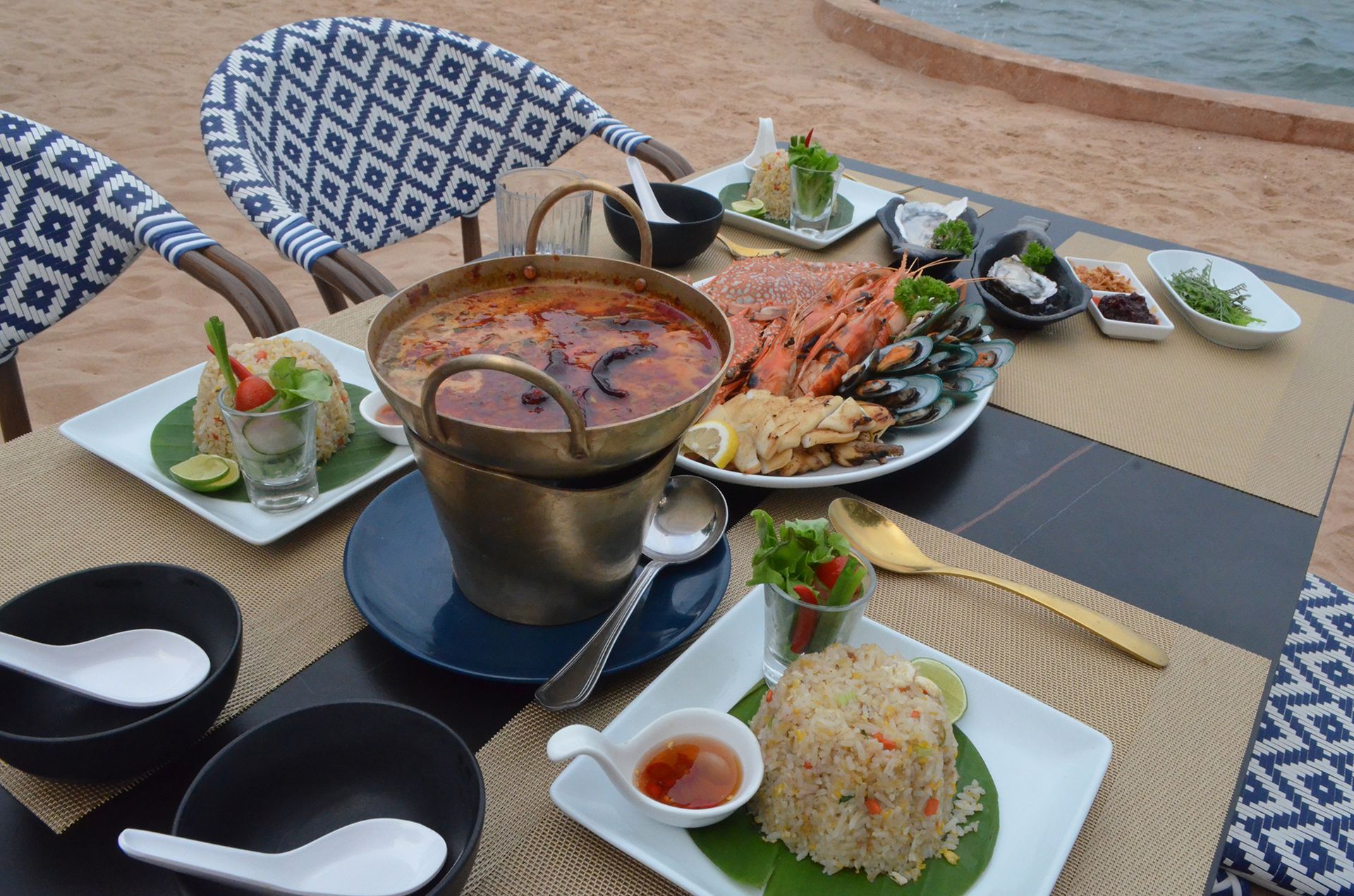 Seafood feast on a beach: Tom Yum soup, fried rice, mussels, and lobster. Table set for dining with ocean view.