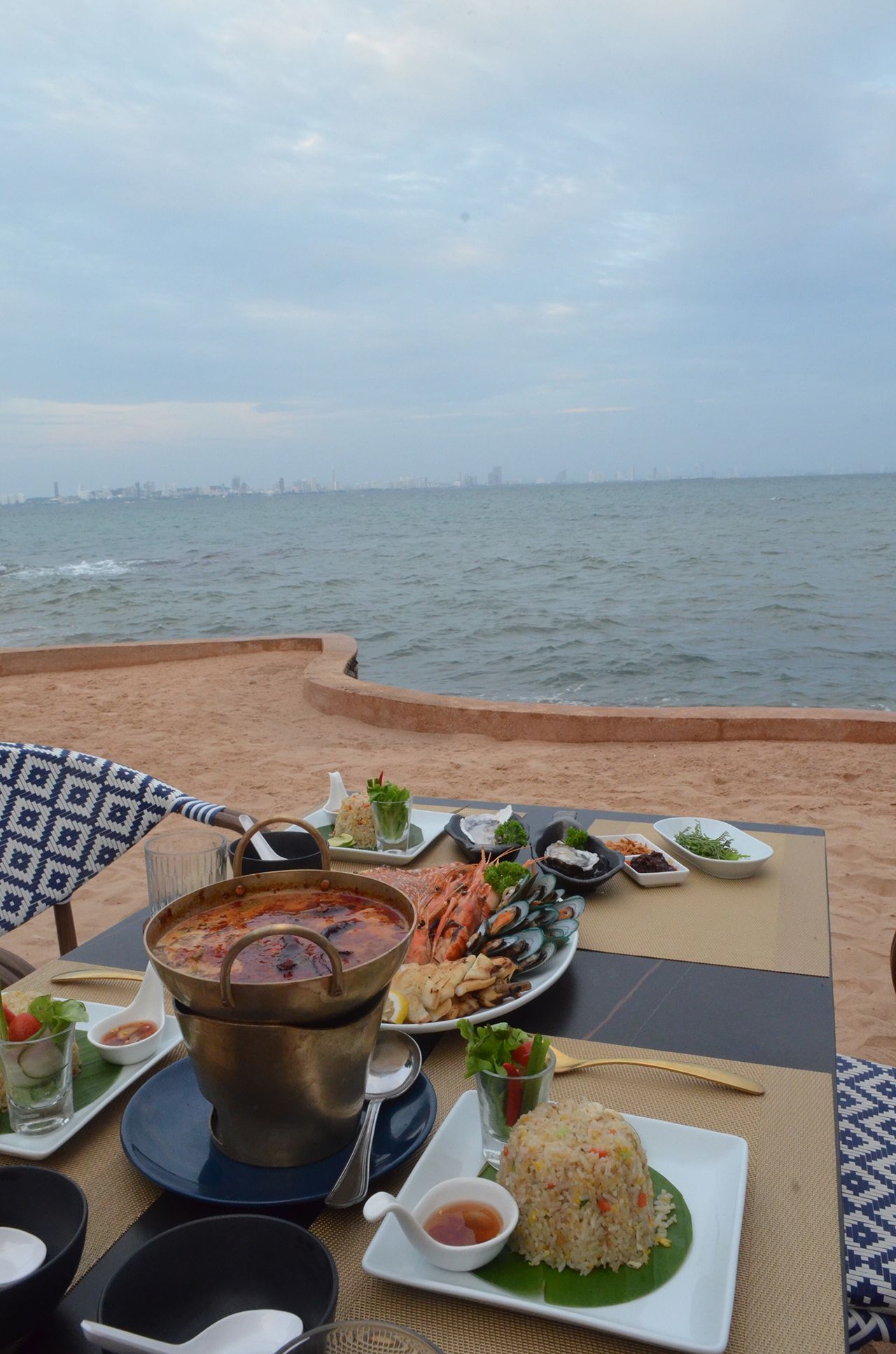 Table of seafood dishes on a beachfront. Blue water, gray sky, and a distant city skyline are in view.