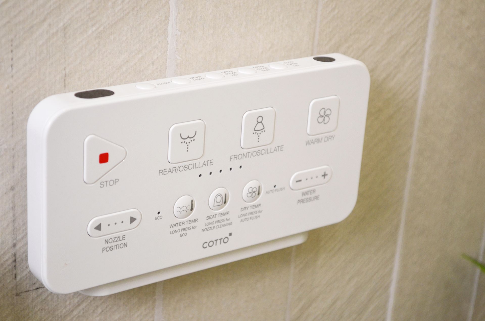 White bidet control panel with buttons for various functions mounted on a tiled wall.