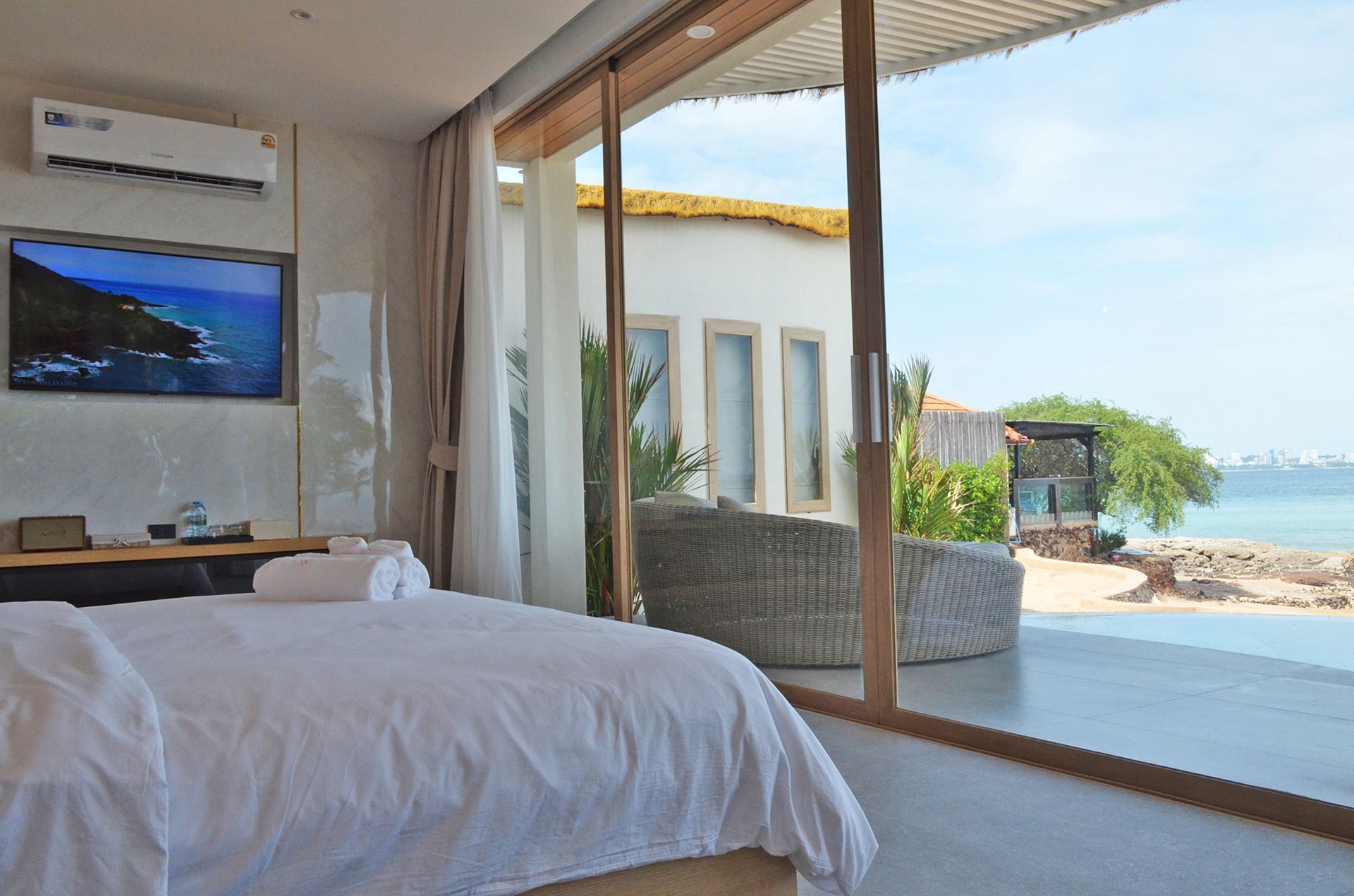 Bedroom with large window overlooking a beach. Bed is white, tv mounted, gray wicker furniture outside.