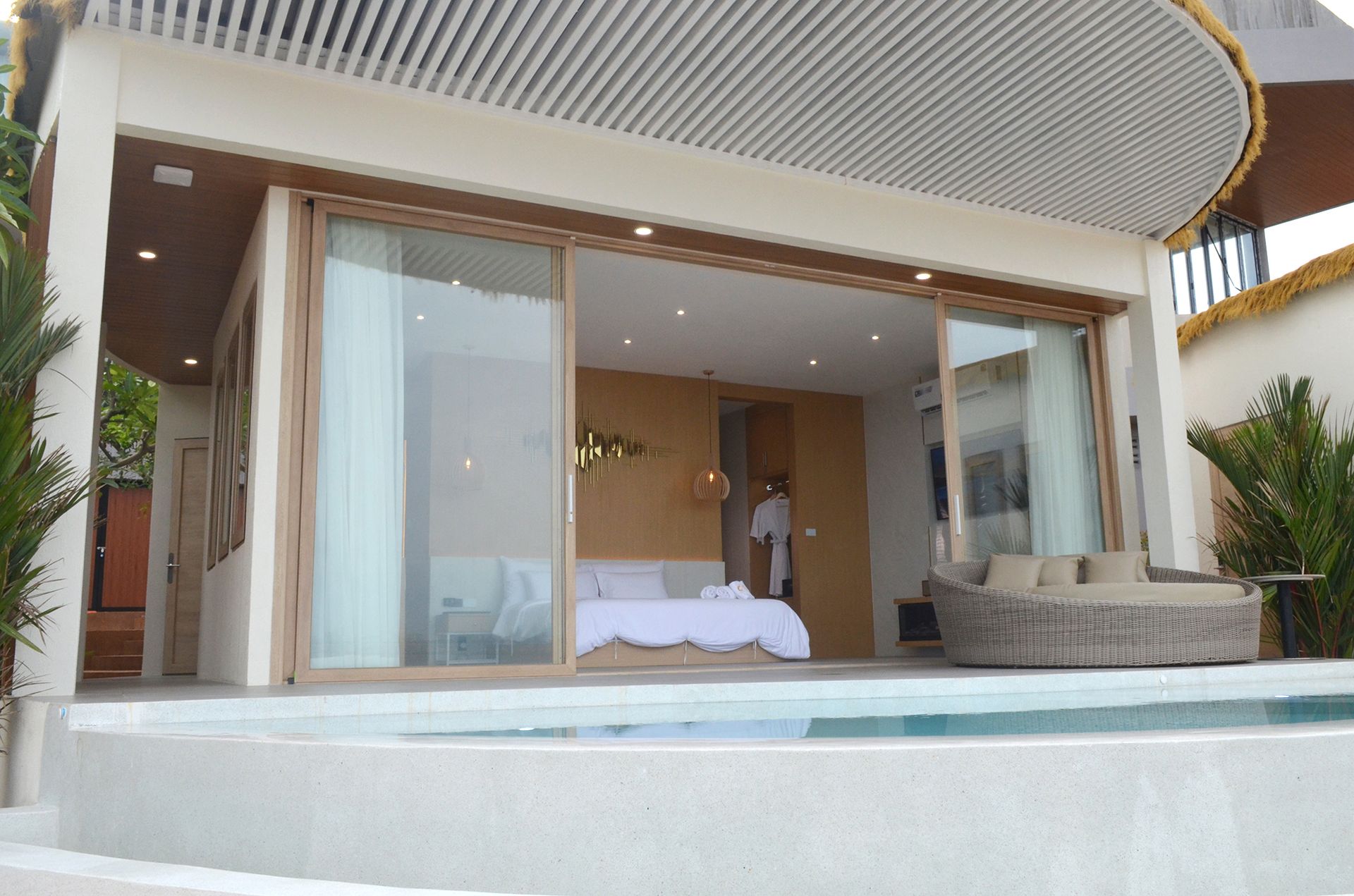 Open-air bedroom with sliding glass doors, pool, and lounge seating. Neutral tones.