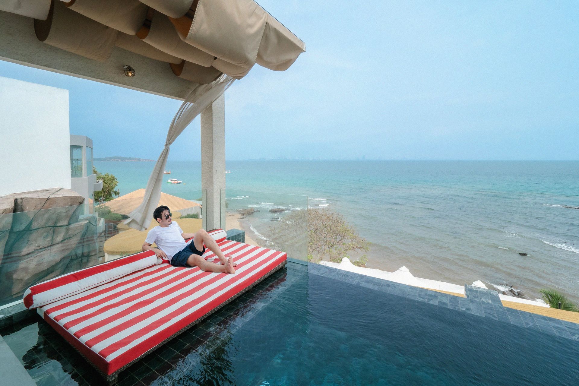Man relaxing on a red and white striped daybed by a pool overlooking the ocean.