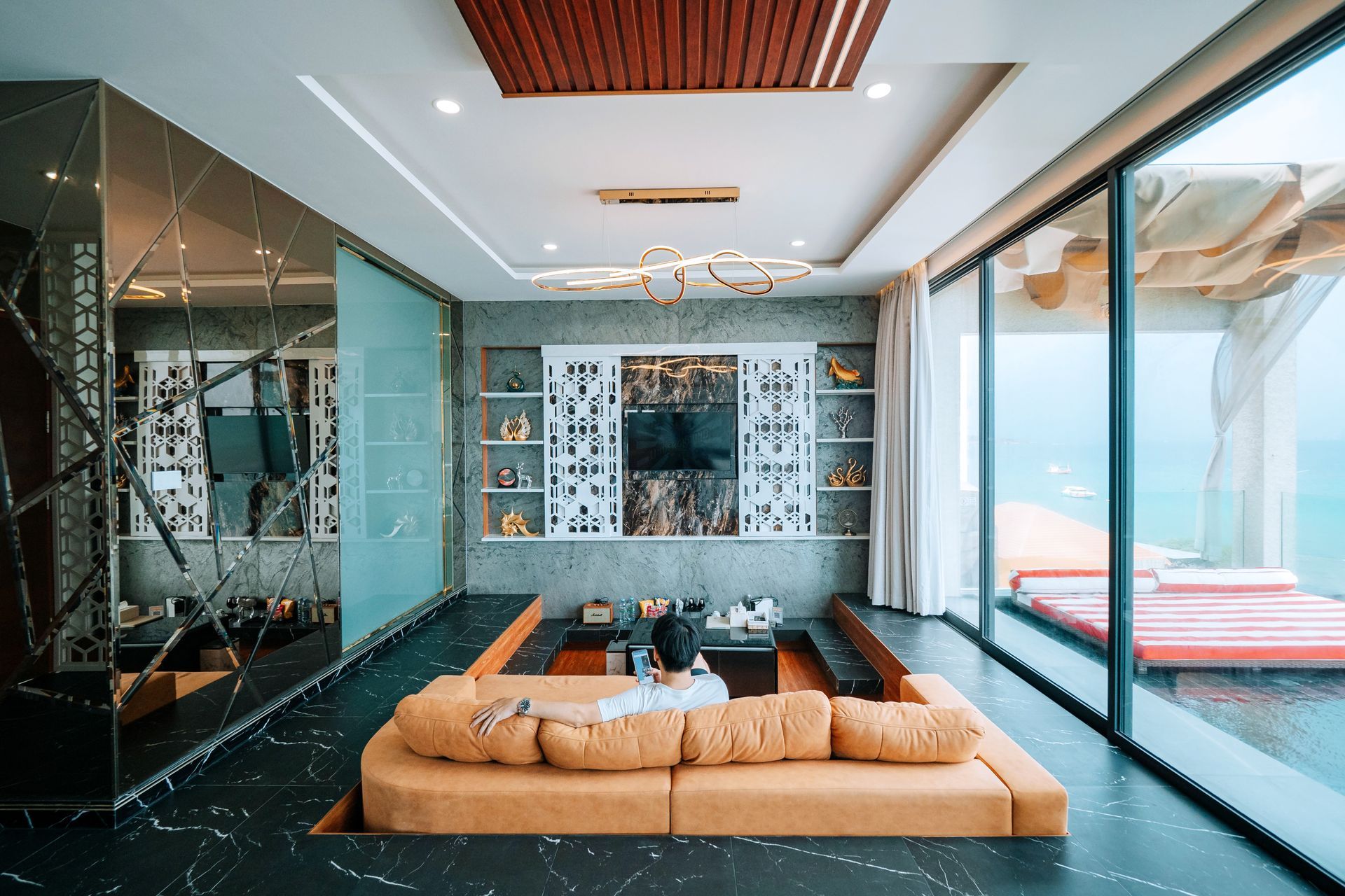 Modern living room with orange sunken sofa, large windows overlooking water, decorative shelves, and marble floor.