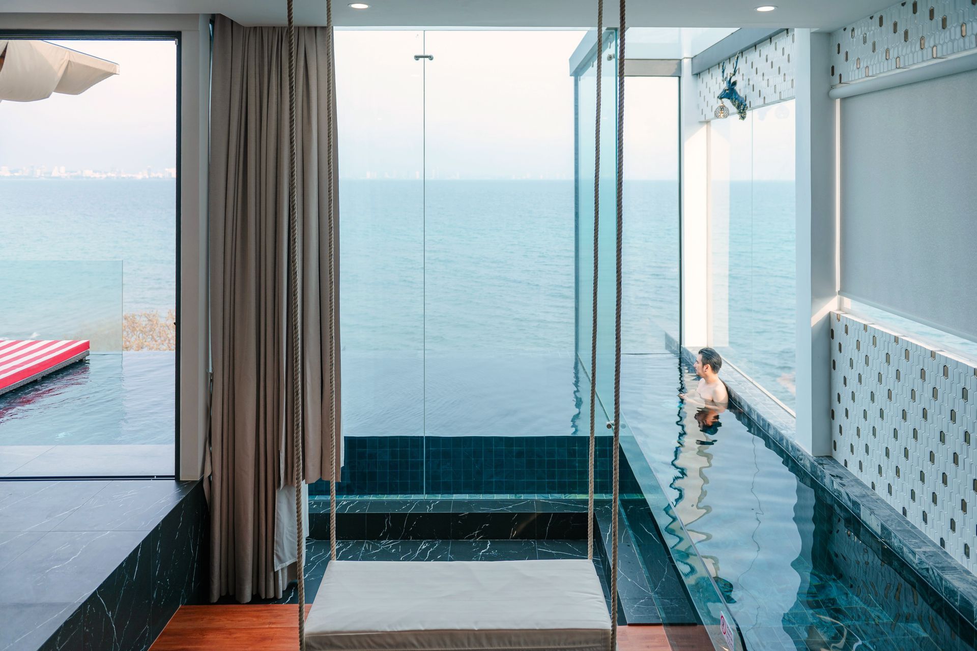 Modern pool with ocean view; person stands in water. White swing, large windows, dark tile.