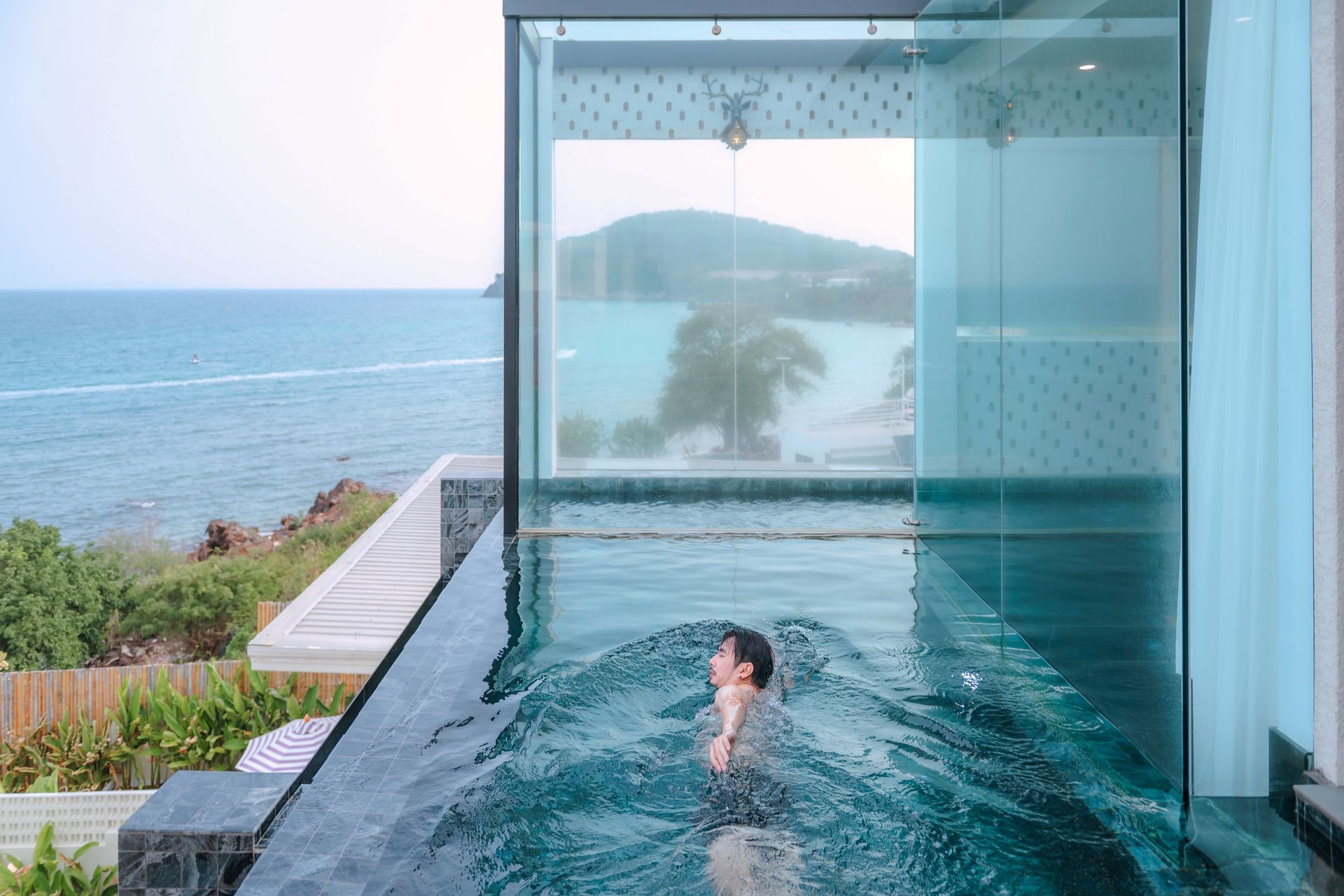 Person swimming in an infinity pool overlooking the ocean, with a modern building and overcast sky.