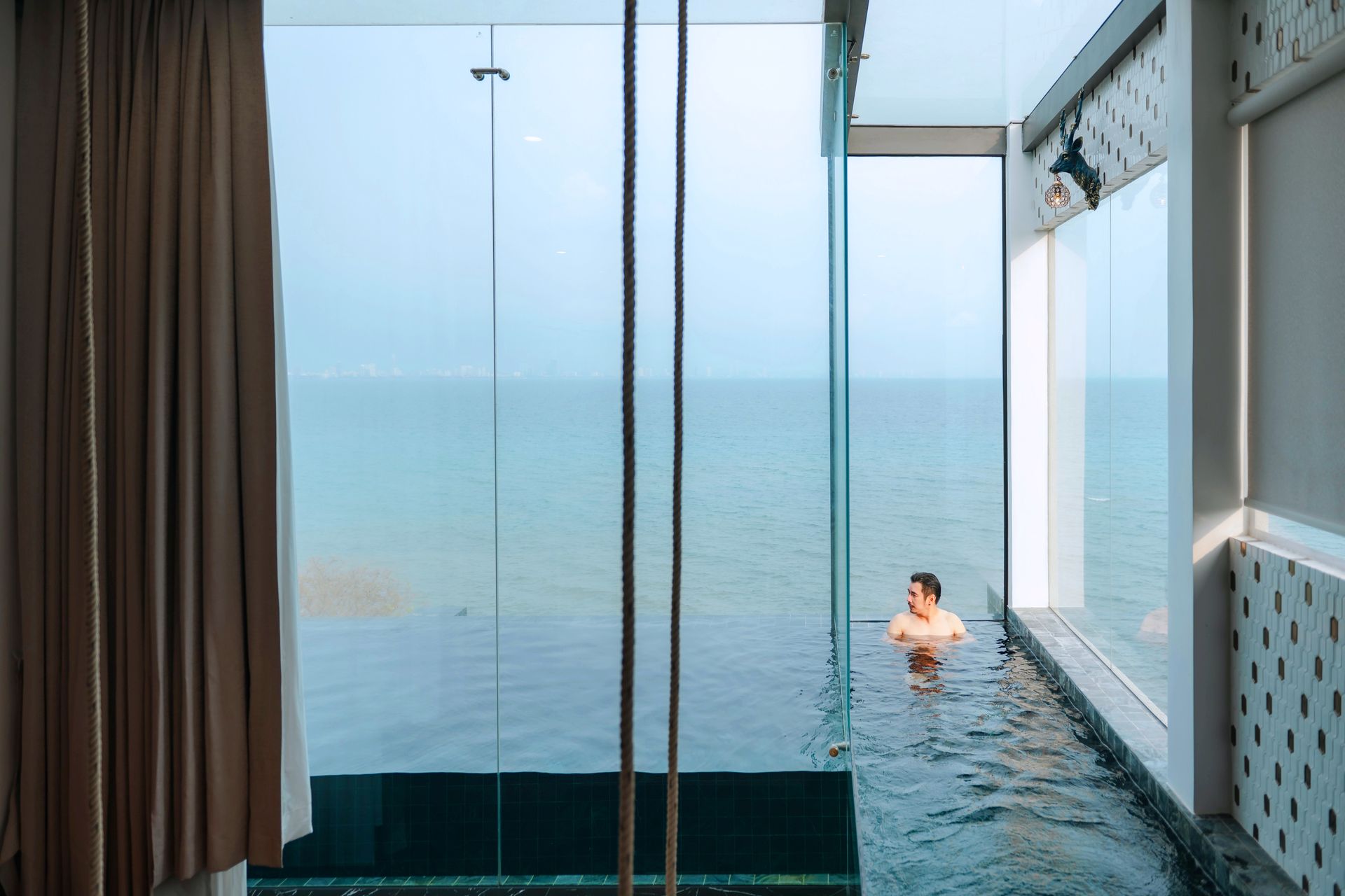 Person in a pool looking at the ocean through a large window. Beige curtain, white walls.