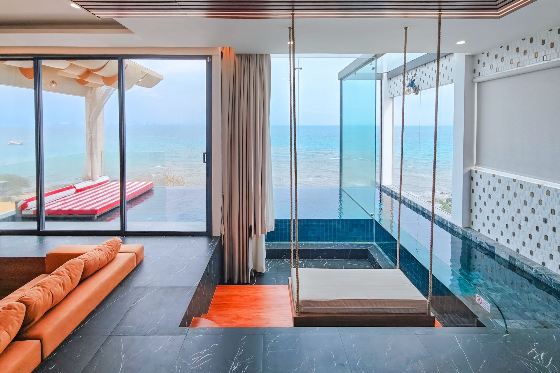 Luxury room with a swing hanging over a pool, overlooking the ocean. Orange couch, large windows, and outdoor cabana.