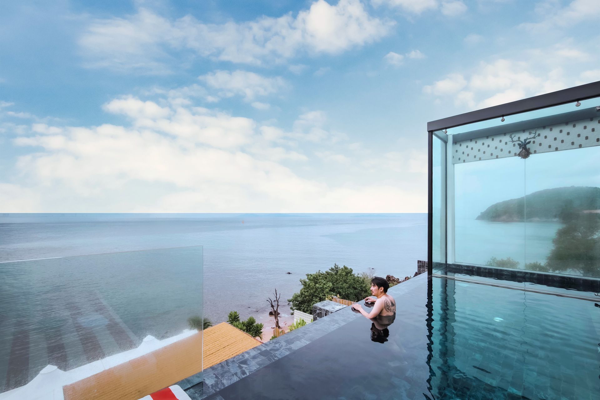 Woman in infinity pool overlooking the ocean, framed by a glass shower wall, under a cloudy blue sky.