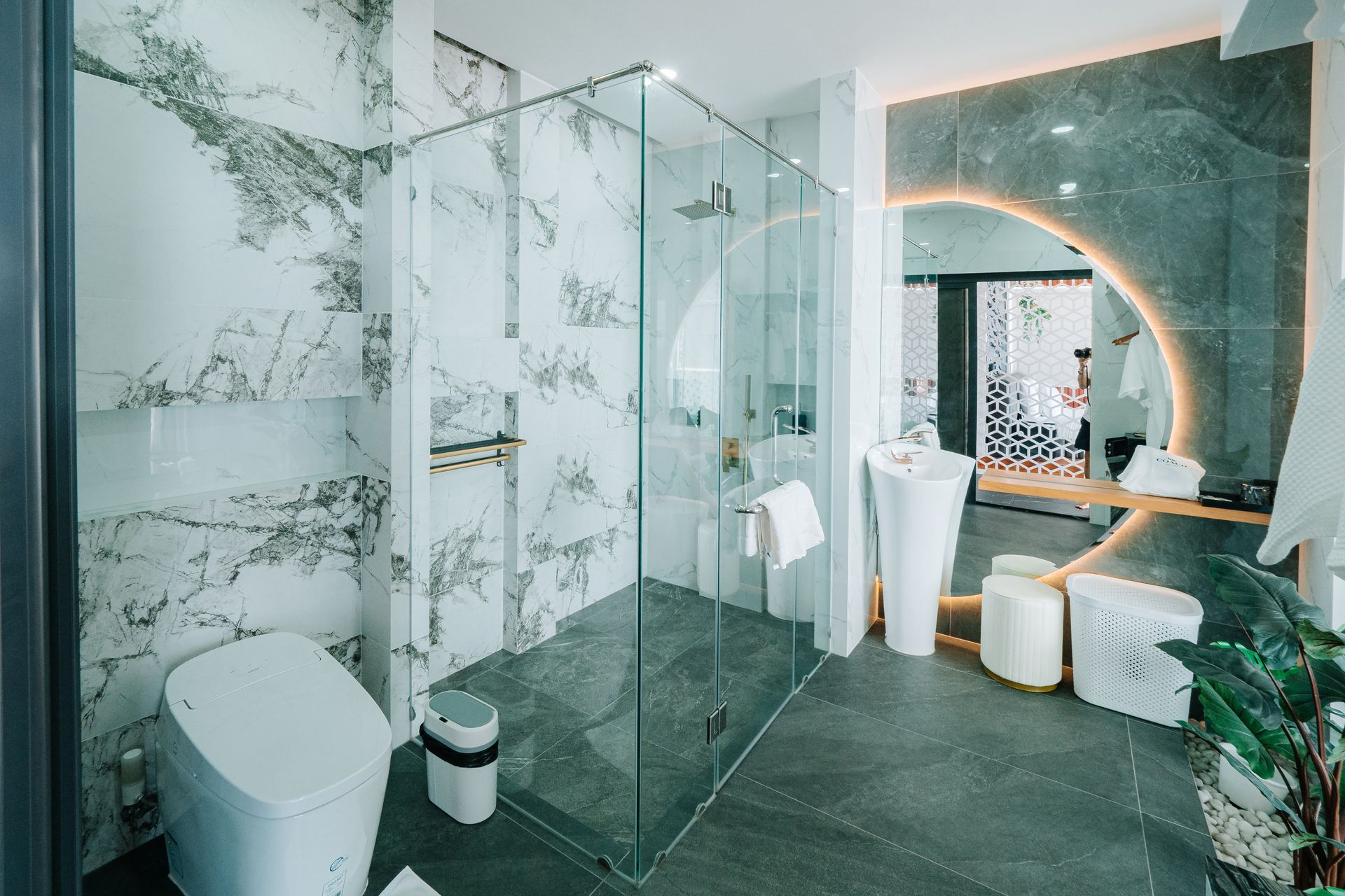 Modern bathroom with marble walls, glass shower, and round mirror with light.