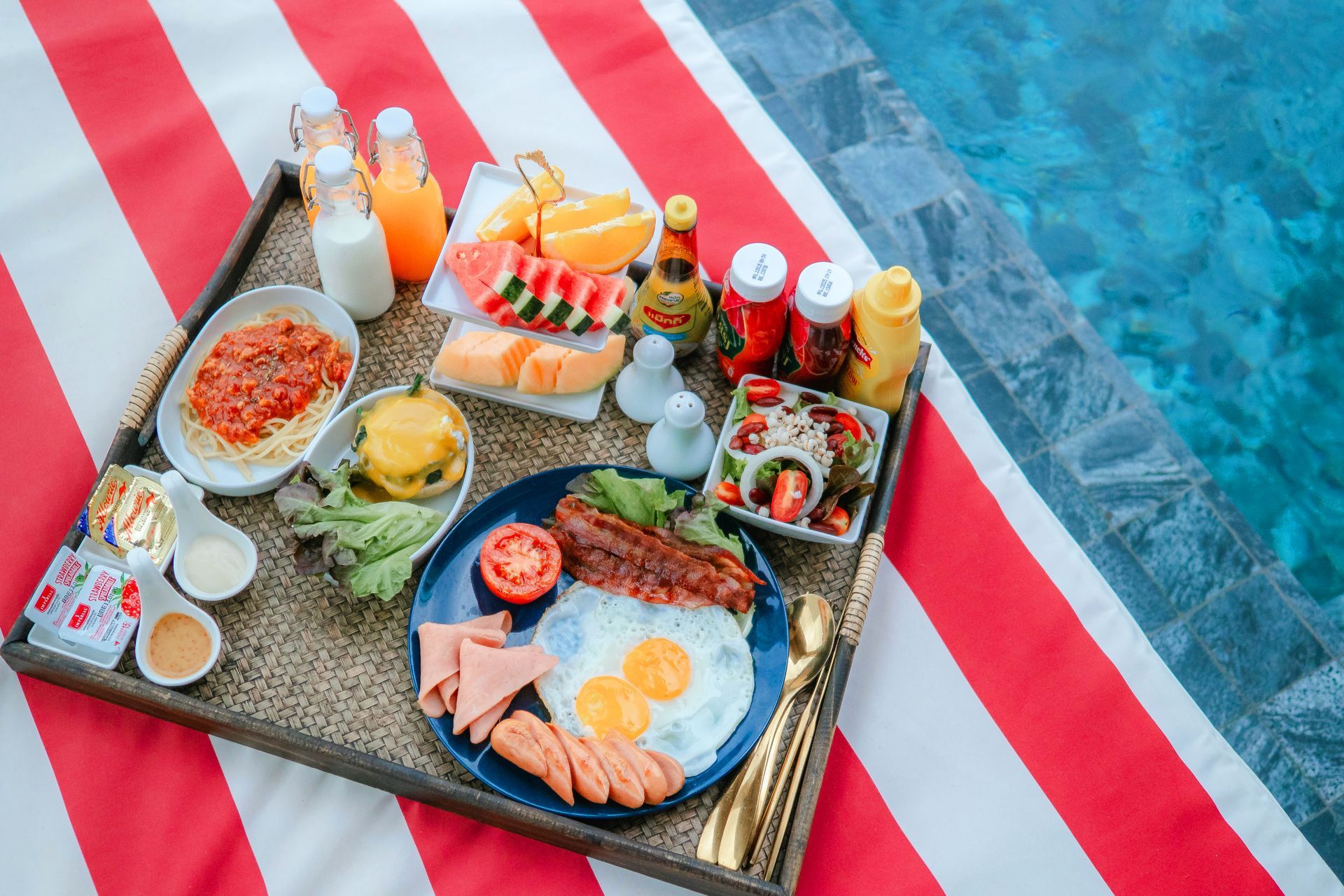 Breakfast tray by a pool with eggs, fruit, and juice on red and white striped fabric.