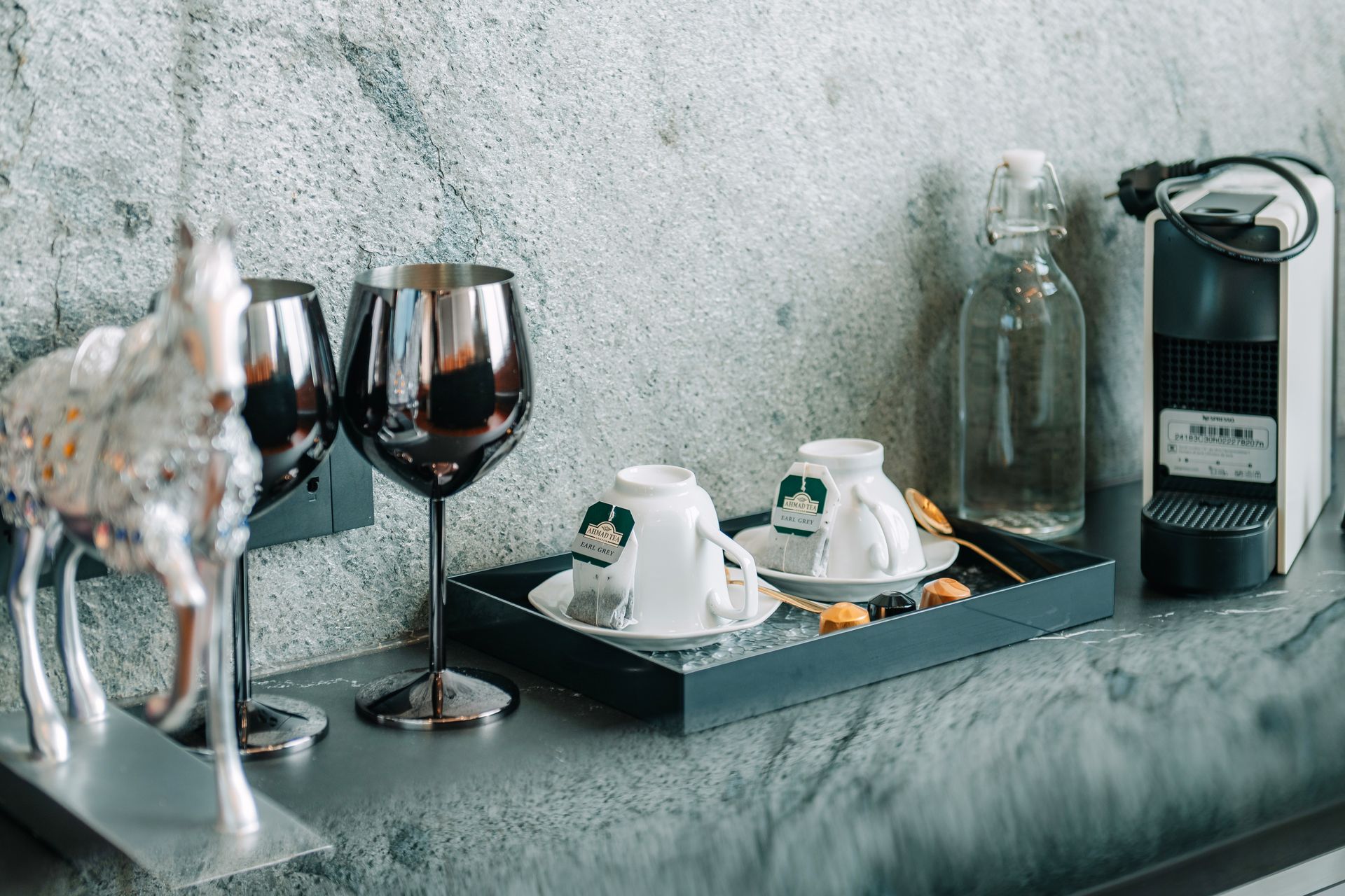 Silver glasses, tea cups, water bottle, and coffee machine on a stone countertop with a decorative horse statue.