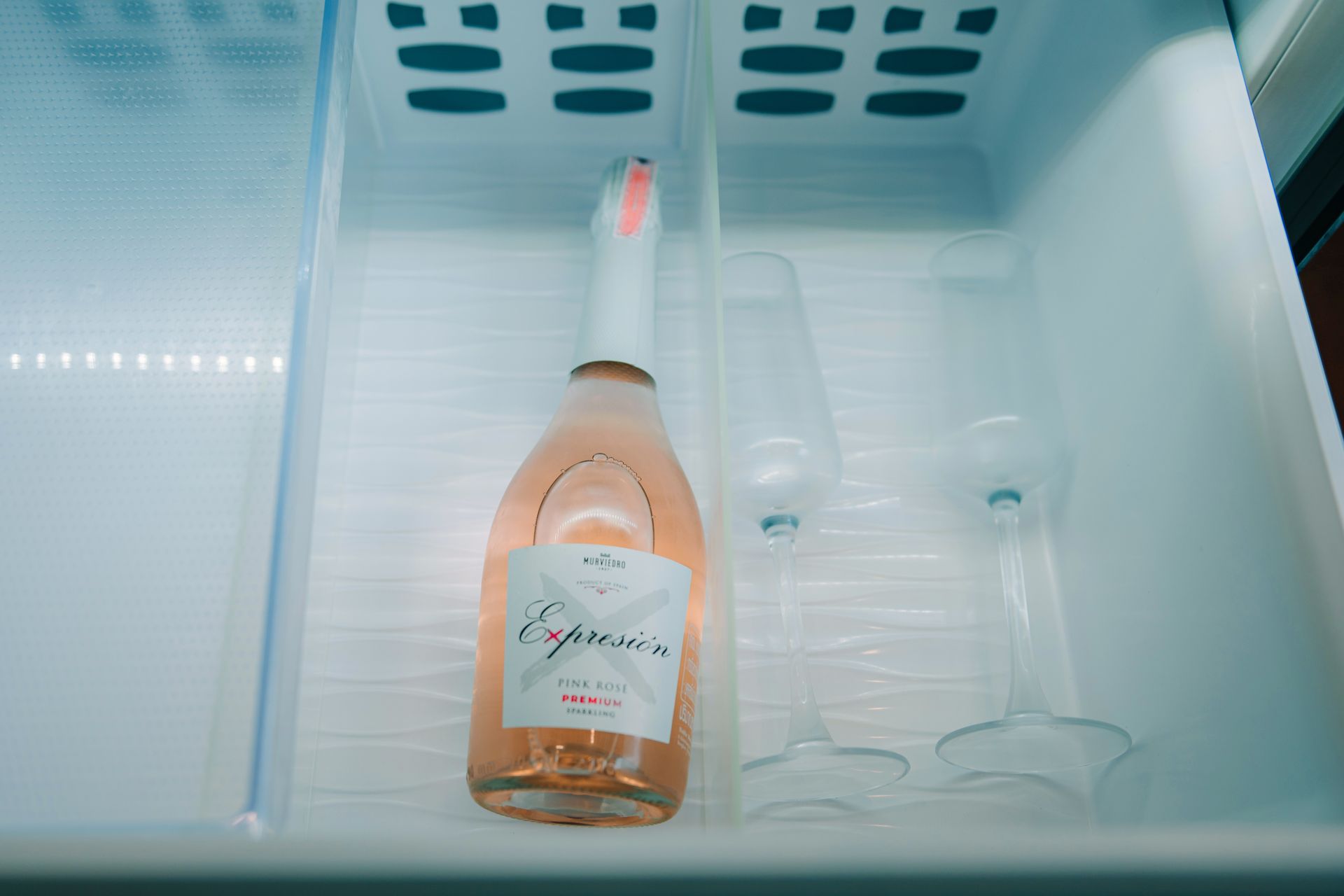 A bottle of rose sparkling wine and two glasses inside a refrigerator drawer.