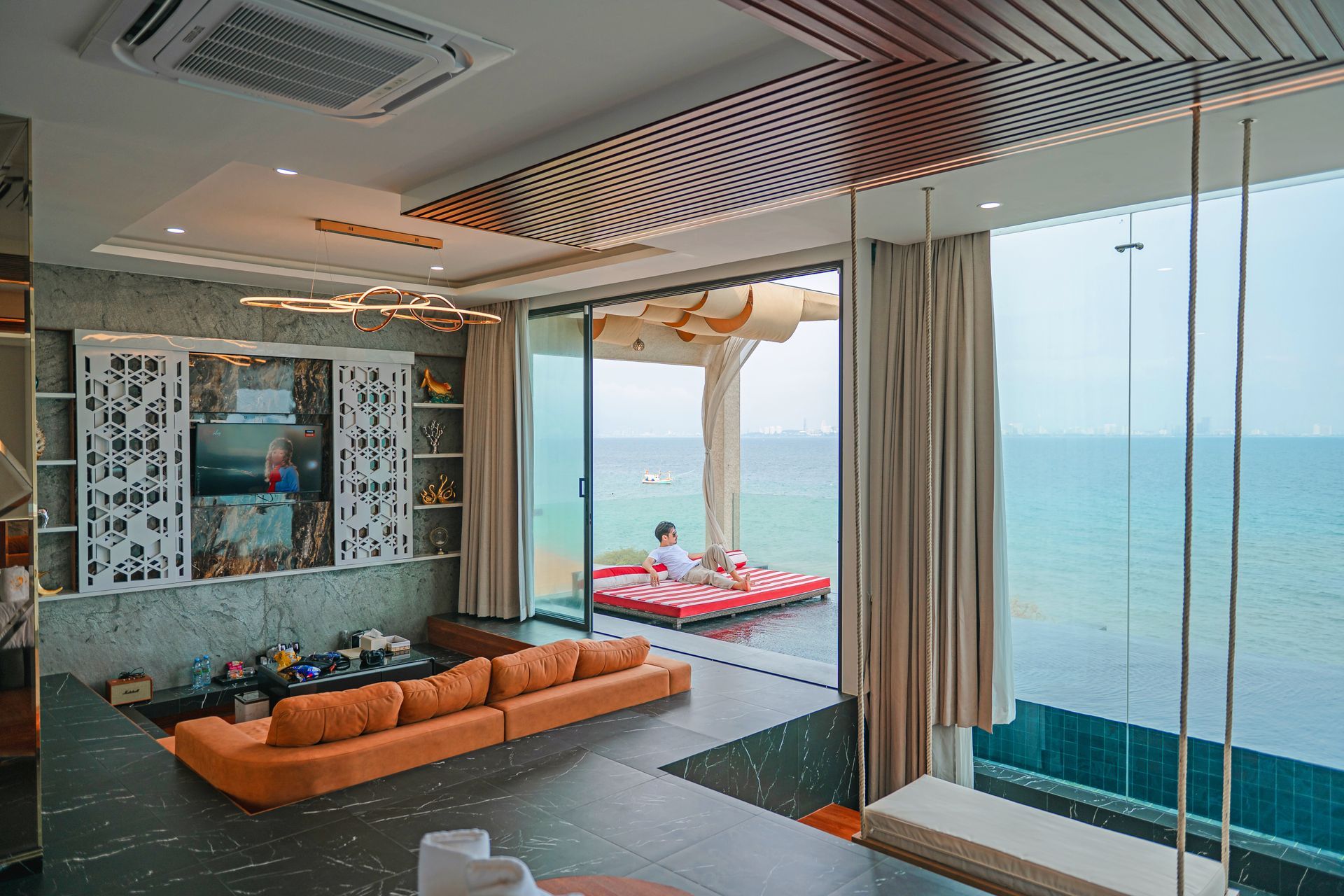 Interior view of luxury seaside home with orange sofa, open to deck over water, person relaxing.