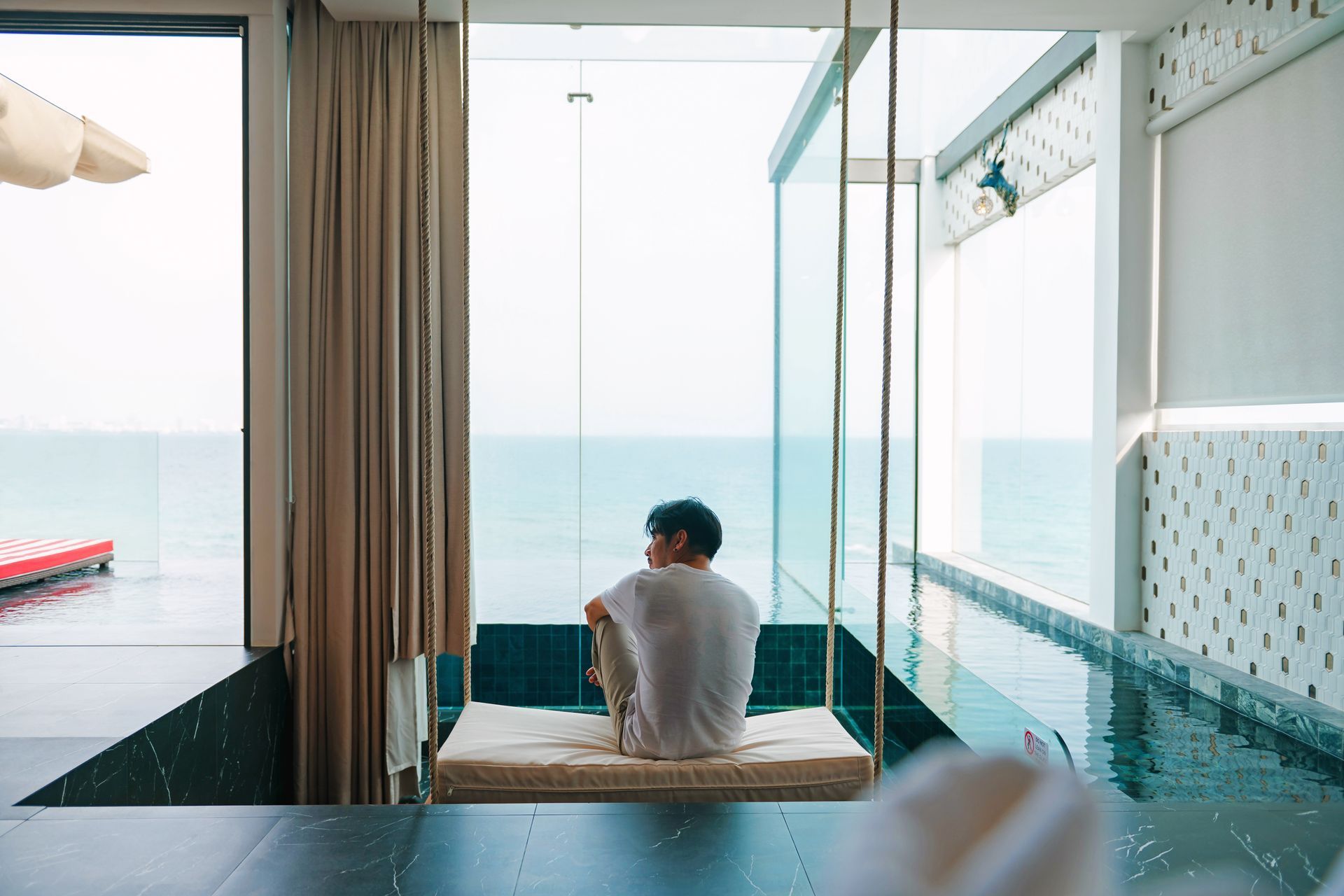 Person sitting on swing looking at the ocean through a window next to a pool.