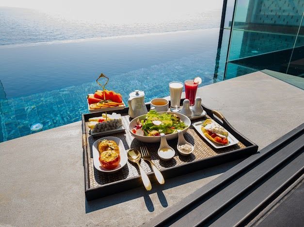 Breakfast tray with fruit, salad, drinks, and other food, by a pool.