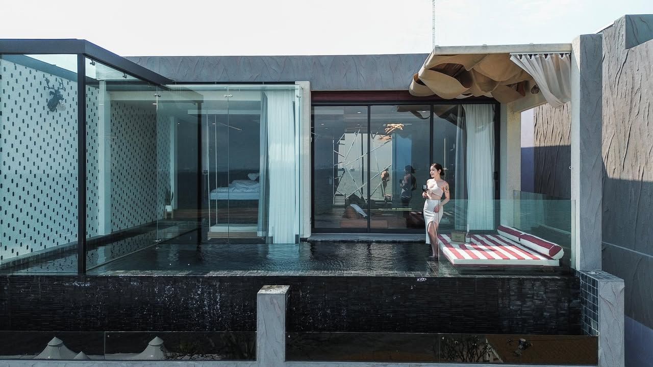 Woman in a white towel stands at the edge of a small pool by a glass-walled room.