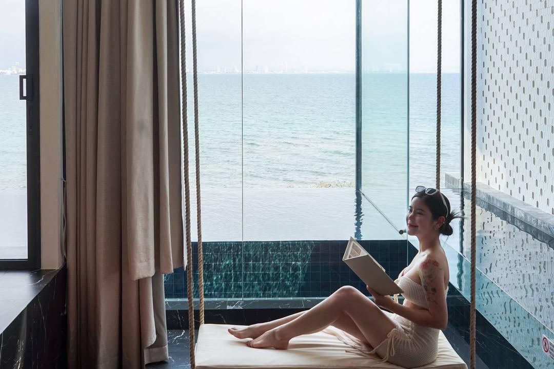 Woman on a swing reading a book, ocean view through a large window.
