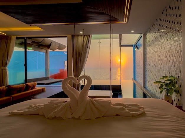Bedroom with swan-shaped towel art, large windows overlooking water, sunset view.