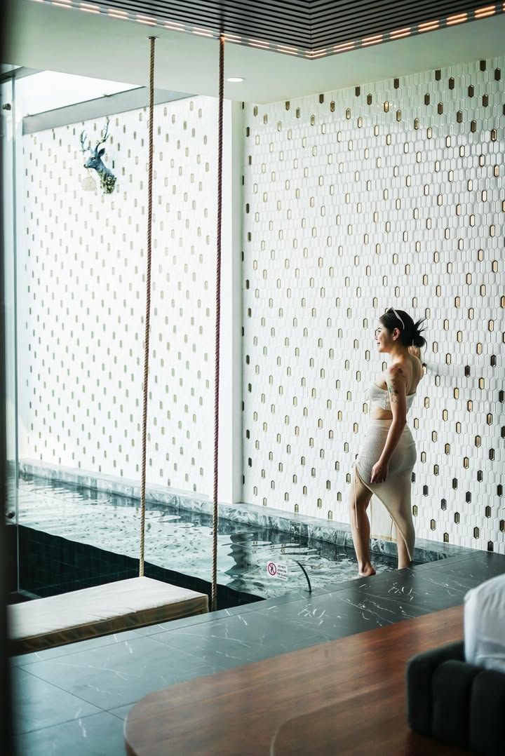 Woman standing in water, gazing at textured wall. Swing hangs nearby. Modern, white interior.