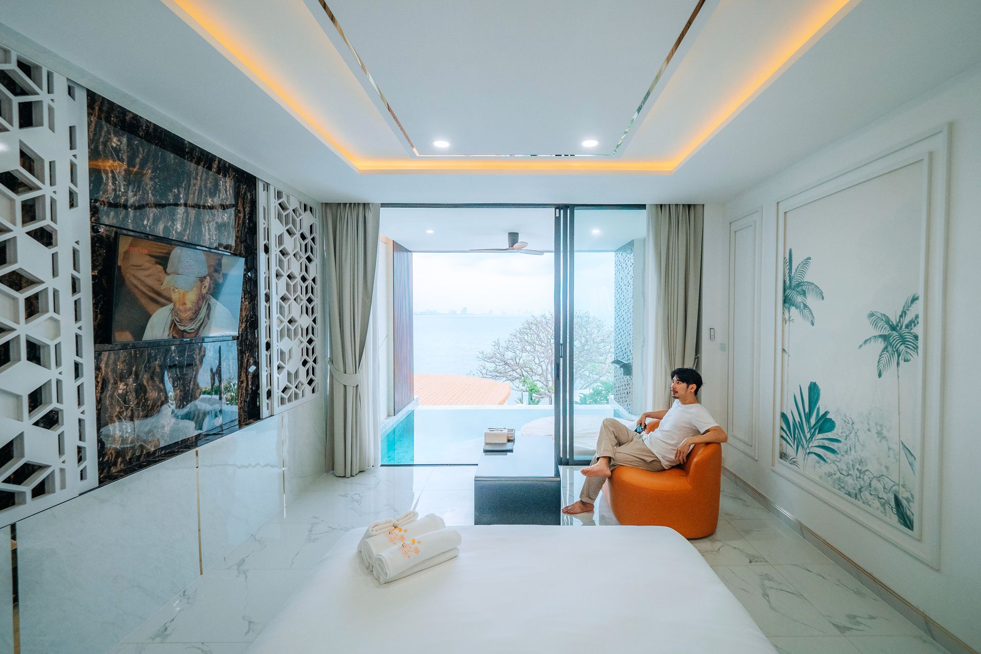 A person sits in an orange chair, looking out at a pool. Modern bedroom with a TV and decorative wall.