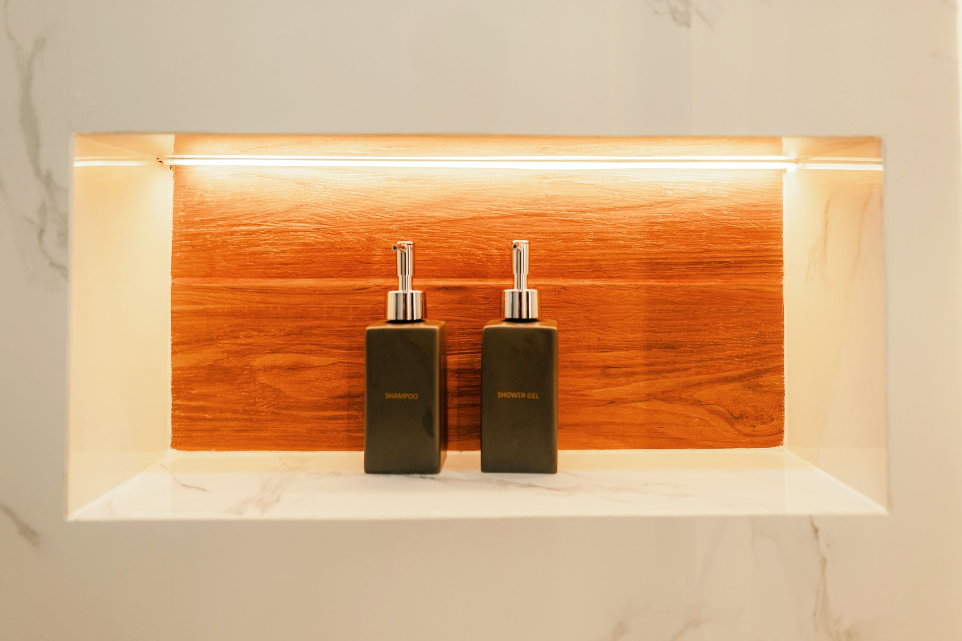 Two dark bottles with silver tops in a recessed, lit shelf with wood backing and white trim.