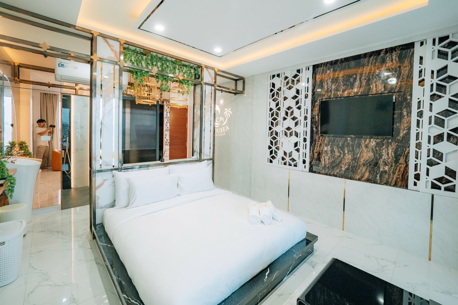Modern hotel bedroom with white bedding, TV, decorative wall panels, and a view into a bathroom.