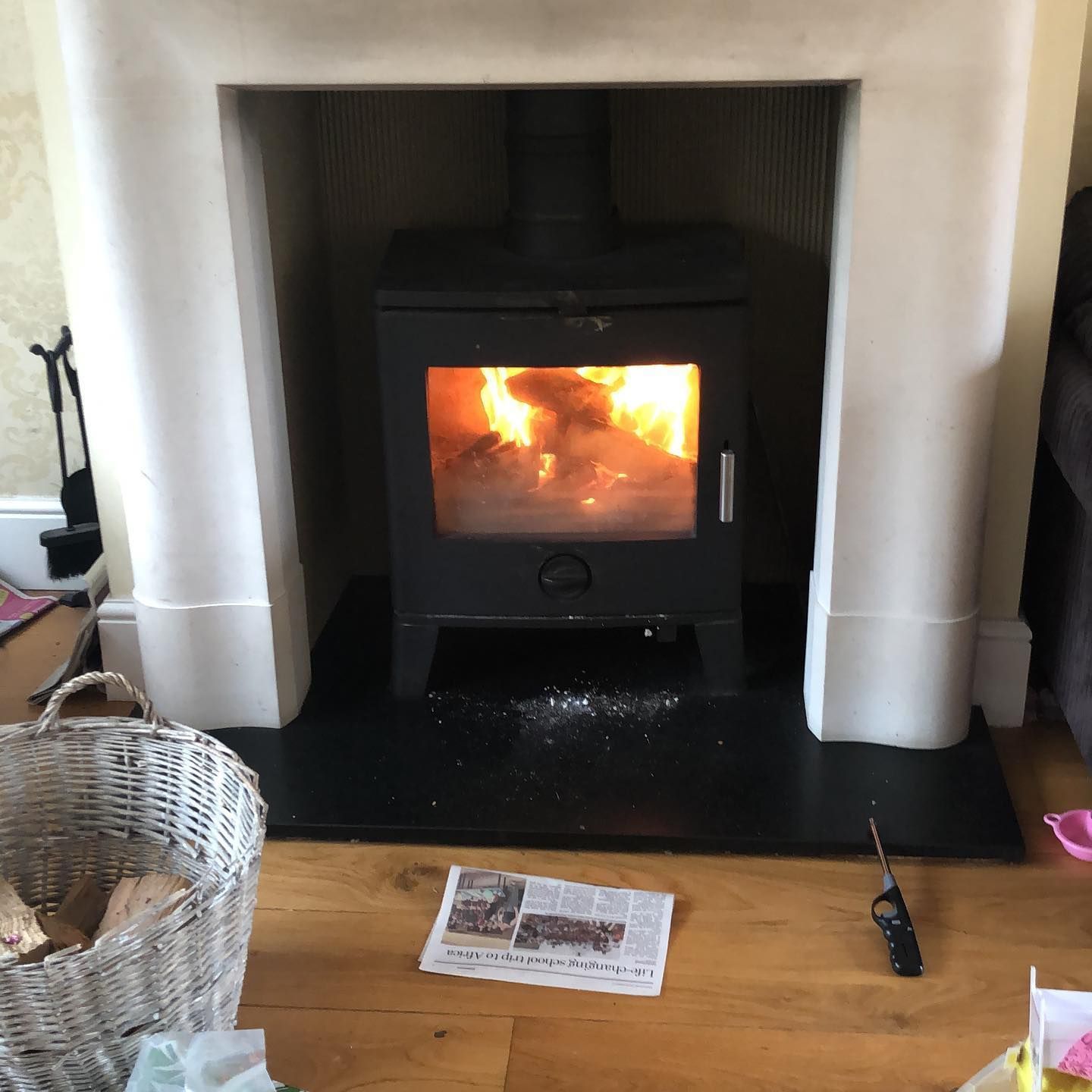 A lit black wood-burning stove inside a white fireplace, with a basket of wood and a newspaper nearby.