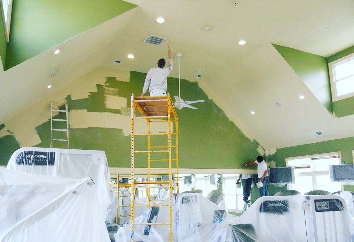 Painters on scaffolding in a room with green walls, painting a beige ceiling. Gym equipment is covered.