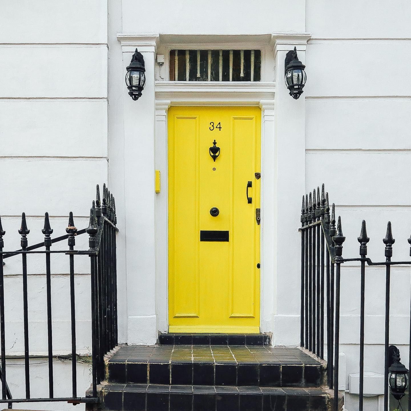 Yellow front door with address number 34 in white, flanked by black sconces on white building, black wrought iron fence.