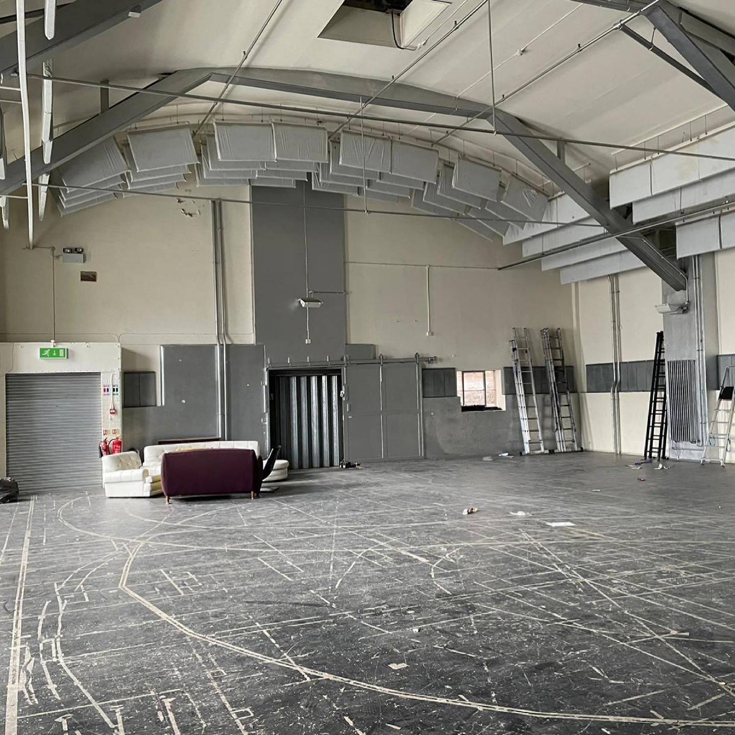 Empty, open-plan room with arched ceiling, gray walls, and distressed black flooring. White couches and equipment are v