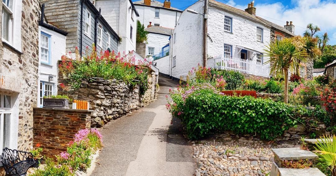 Narrow cobblestone street in a quaint village, lined with stone walls and vibrant flowers, white buildings.